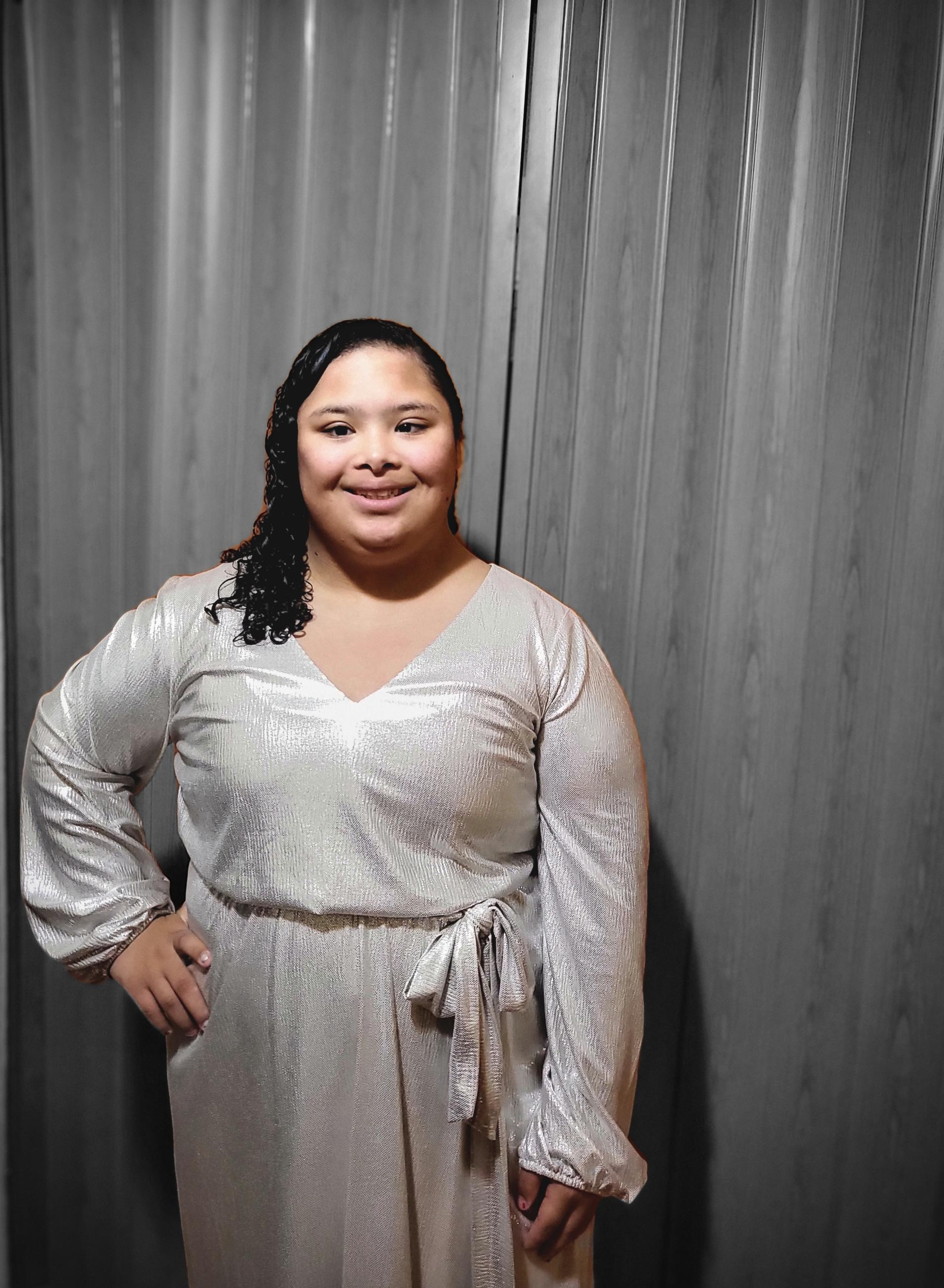 woman with down syndrome with dark brown hair and brown eyes smiling at the camera in her silver dress, one hand on one hip