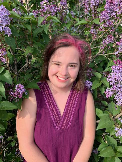 Woman with down syndrome and brown hair is surrounded by flowers as she smiles at the camera