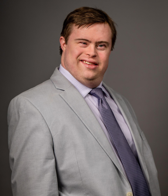 Adrian, a white man with Down syndrome, poses for a headshot. He has brown hair and is wearing a white collared shirt, tan suit jacket, and a muted purple tie.